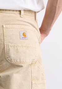 Tan cotton work pants with a rectangular back pocket featuring a white patch and yellow logo. Visible stitching detail.