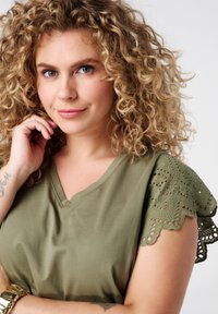 T-shirt vert olive avec col en V, présentant des accents de dentelle sur les manches courtes. Texture douce en coton. Cheveux bouclés, main touchant doucement le visage.
