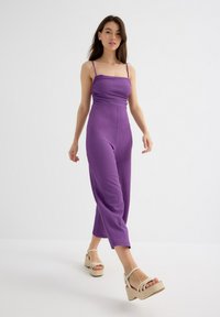 Mono morado acanalado con tirantes finos, cuerpo ajustado y piernas anchas. La modelo lleva sandalias de plataforma beiges con correas en el tobillo. Fondo liso.