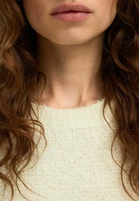 Jasny, textured sweter, zbliżenie na szyję i usta osoby, z falującymi brązowymi włosami otaczającymi twarz.