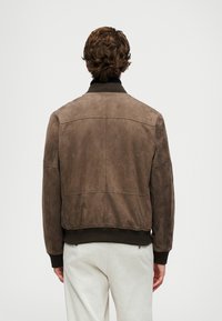 Man wearing brown suede bomber jacket and light-colored pants, standing facing away against plain light background.