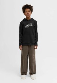 Hoodie nero con logo frontale, tasca a marsupio e polsini a costine, abbinato a pantaloni di velluto a coste marroni dal taglio regolare e sneakers bianche.