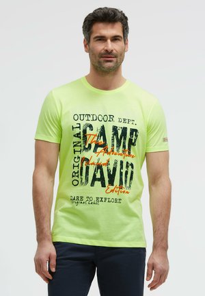 Mann trägt ein hellgrünes T-Shirt mit schwarzem und orangem Text, steht vor einem einfarbigen hellen Hintergrund, schaut nach vorne mit neutralem Gesichtsausdruck.