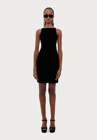 Rochie din catifea neagră cu o siluetă ajustată, decolteu pătrat și lungime deasupra genunchiului, asortată cu sandale negre cu toc înalt.