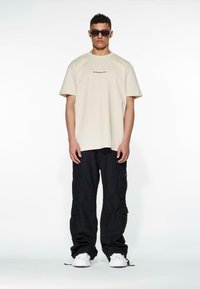 Beige oversized t-shirt with logo, paired with black cargo pants featuring pockets, complemented by white sneakers and dark sunglasses.