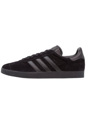 Czarny zamszowy niski sneaker Adidas z czarnymi skórzanymi paskami, czarną teksturowaną podeszwą i sznurowanym zamknięciem, pokazany z profilu.