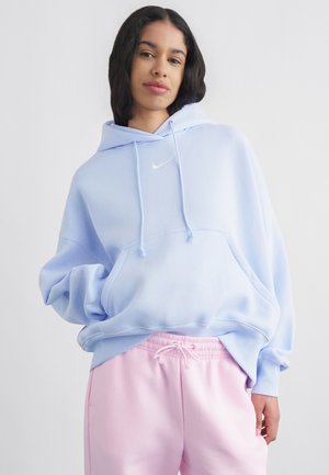 Νέο άτομο με ανοιχτό μπλε φούτερ Nike με κουκούλα και ροζ παντελόνια με κορδόνι, στέκεται με το ένα χέρι στην τσέπη μπροστά από απλό φόντο.