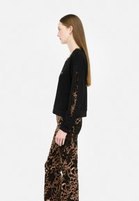 Donna di profilo che indossa un top lungo nero con maniche a taglio sopra pantaloni larghi a stampa leopardo, su uno sfondo bianco.