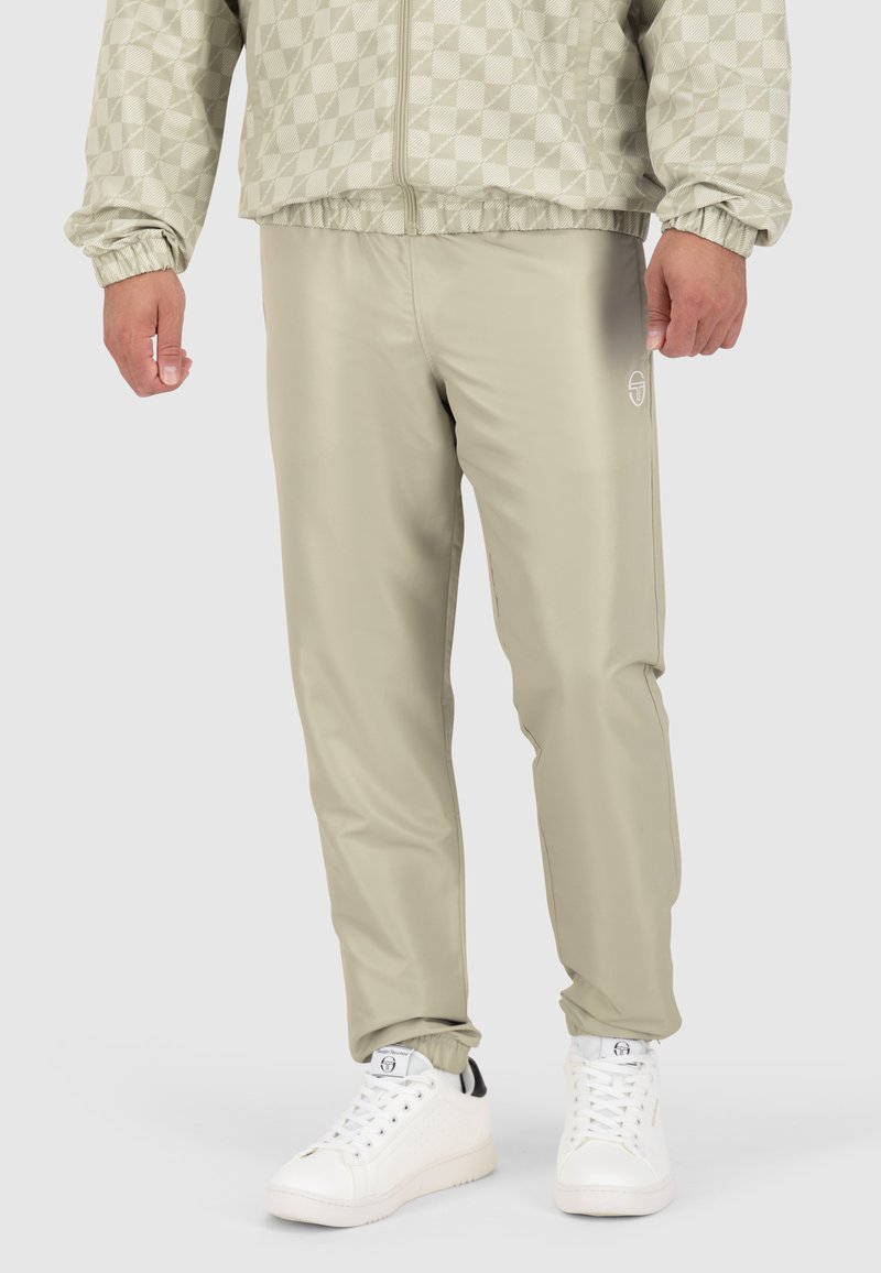 Helle beige Sweatpants mit elastischem Bund, taillierter Passform und kleinem Logo an der Seite; kombiniert mit weißen Turnschuhen. Glatte Textur.
