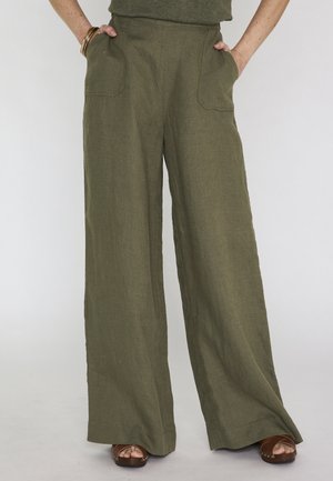 Donna che indossa pantaloni a gamba larga verde oliva con tasche frontali e sandali platform marroni, in piedi con le mani nelle tasche.