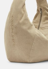 Beżowa torba z materiału o miękkiej, zaokrąglonej sylwetce. Posiada boczny zamek błyskawiczny oraz tłoczone napisy "LIEBESKIND BERLIN" jako wyróżniający się detal.