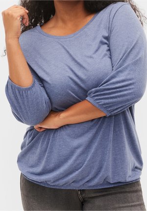 Femme portant une chemise bleue à manches longues avec des poignets élastiques, associée à un pantalon gris foncé, posant les bras croisés et la main près du visage.