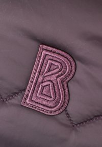 Vijolična tkanina s strukturirano, vezeno oznako "B" v temnejši vijolični barvi, ki vključuje geometrijske oblike in natančno šivanje.