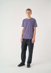Lila Baumwoll-T-Shirt mit Rundhalsausschnitt, kurzen Ärmeln und kleinem Logo auf der Brust, kombiniert mit dunklen, lässig sitzenden Hosen und schwarzen Schuhen.