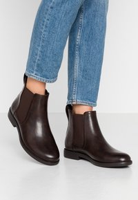 Botas de cuero marrón hasta el tobillo con paneles laterales elásticos y punteras redondeadas. Suela resistente, textura suave, combinadas con jeans azul claro con puños.