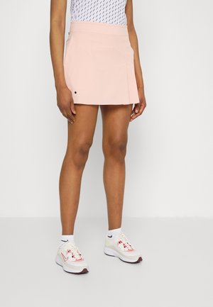 Persoon draagt een lichtroze minirok, een wit patroon topje, witte sokken en witte sneakers met roze accenten, staand op een witte vloer.