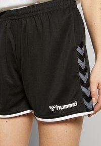Svarta sportshorts med vit trim, chevronränder på sidan och en vit Hummel-logotyp nära fållen, bärs av en person.