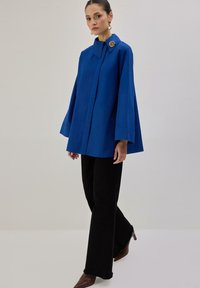 Modro velika tunika z ovratnikom in okroglim okrasnim gumbom. Kombinirana s črnimi razširjenimi hlačami in rjavimi škornji z visokimi petami. Gladek tekstura.