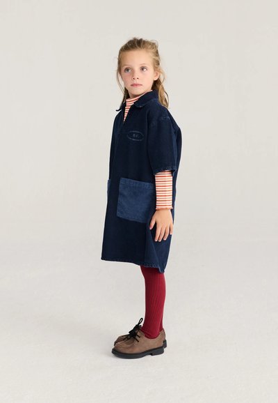 Jeune fille debout portant une robe bleu marine avec des poches, des manches longues rayées rouges, des collants rouges et des chaussures à lacets marron sur un fond uni.