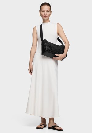 Donna che indossa un abito lungo bianco senza maniche, sandali neri, orecchini a cerchio dorati e tiene una borsa a tracolla nera con trama e con una tracolla larga.