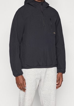 Mann trägt eine schwarze Kapuzenpulloverjacke mit Reißverschlusstasche auf der Brust und hellgraue Jogginghose, sichtbar ab der Taille.