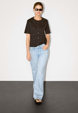 Schwarzes Kurzarm-T-Shirt mit Nieten, kombiniert mit hellblauen weit geschnittenen Jeans und schwarzen Flip-Flops, posierend vor einem schlichten Hintergrund.