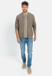Bruine geribde zip-up cardigan over een groene t-shirt, gecombineerd met lichtblauwe spijkerbroek en beige sneakers. Eenvoudig ontwerp met textuuraccenten.