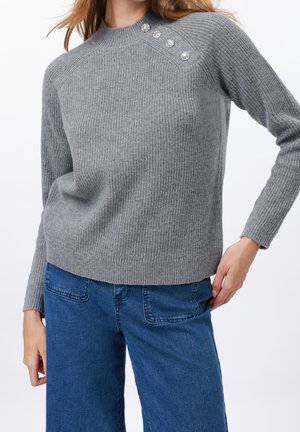 Femme portant un pull en maille côtelée gris avec trois boutons décoratifs sur l'épaule et un jean en denim bleu.
