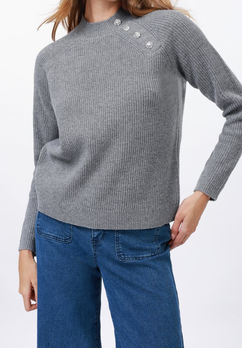 Femme portant un pull en maille côtelée gris avec trois boutons décoratifs sur l'épaule et un jean en denim bleu.
