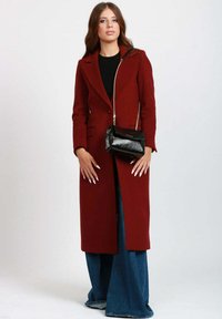 Lungo cappotto bordeaux con revers a punta, chiusura a un bottone e tasche. Indossato sopra un top nero e abbinato a pantaloni in denim a gamba larga e una borsa nera.