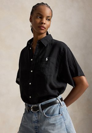 Žena s vrkočmi, oblečená v čiernej krátkorukáve košeli s gombíkmi a logom, svetlomodrých džínsoch, čiernom opasku a náušniciach v tvare mušlí, pozerajúca sa do strany.