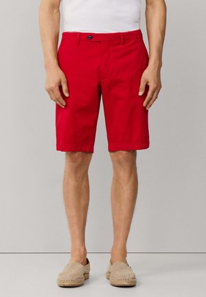 Mann trägt leuchtend rote knielange Shorts, weißes Hemd und beige Slipper, steht vor einem einfarbigen hellgrauen Hintergrund.