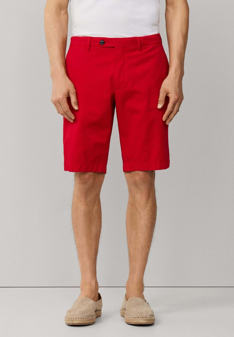 Uomo che indossa pantaloncini rossi vivaci fino al ginocchio, camicia bianca e scarpe beige slip-on, in piedi davanti a uno sfondo grigio chiaro uniforme.