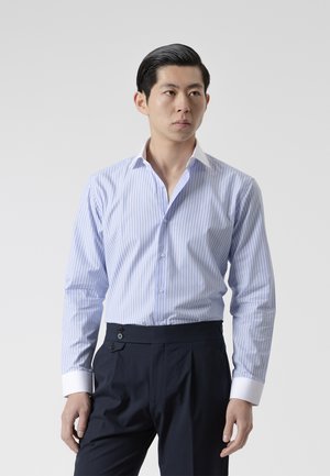 Hombre que lleva una camisa de vestir de rayas azul claro y blanca con puños blancos y pantalones azul marino oscuro, de pie frente a un fondo liso.