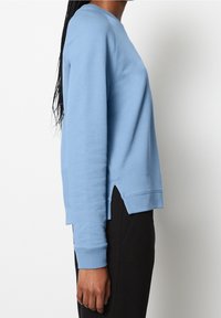 Sudadera azul claro con cuello redondo, mangas largas y aberturas laterales. Hecha de tela suave, con puños y bajo acanalados.