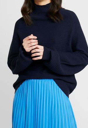 Mujer con suéter de cuello alto azul marino de canalé y falda plisada azul brillante, con las manos entrelazadas cerca del pecho.