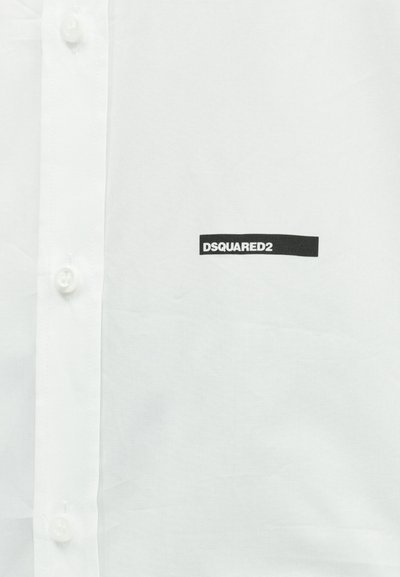 Hvid skjorte med knapper og et sort rektangulært logoemblem med teksten "DSQUARED2" på brystet nær knapperne.