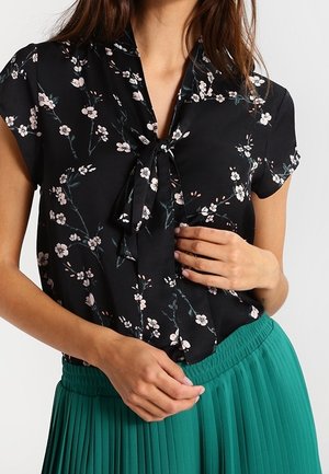 Frau trägt eine schwarze Kurzarmbluse mit kleinem rosa Blumenmuster und gebundener Schleife am Kragen, kombiniert mit einem grünen plissierten Rock.