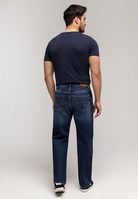 Mężczyzna stojący tyłem, ubrany w granatową koszulkę, ciemnoniebieskie dżinsy z tylnymi kieszeniami oraz czarno-białe trampki na jednolitym tle.