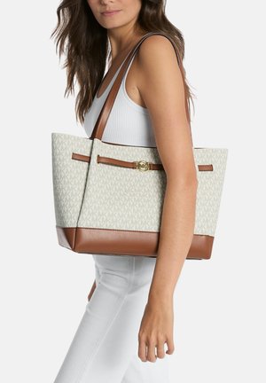 Donna che indossa canotta bianca e pantaloni tiene una borsa beige con rifiniture in pelle marrone e motivo monogramma MK ripetuto.