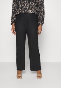 Pantalones plisados negros de cintura alta y corte holgado, combinados con una blusa estampada. La modelo lleva zapatos de tacón abiertos color beige.