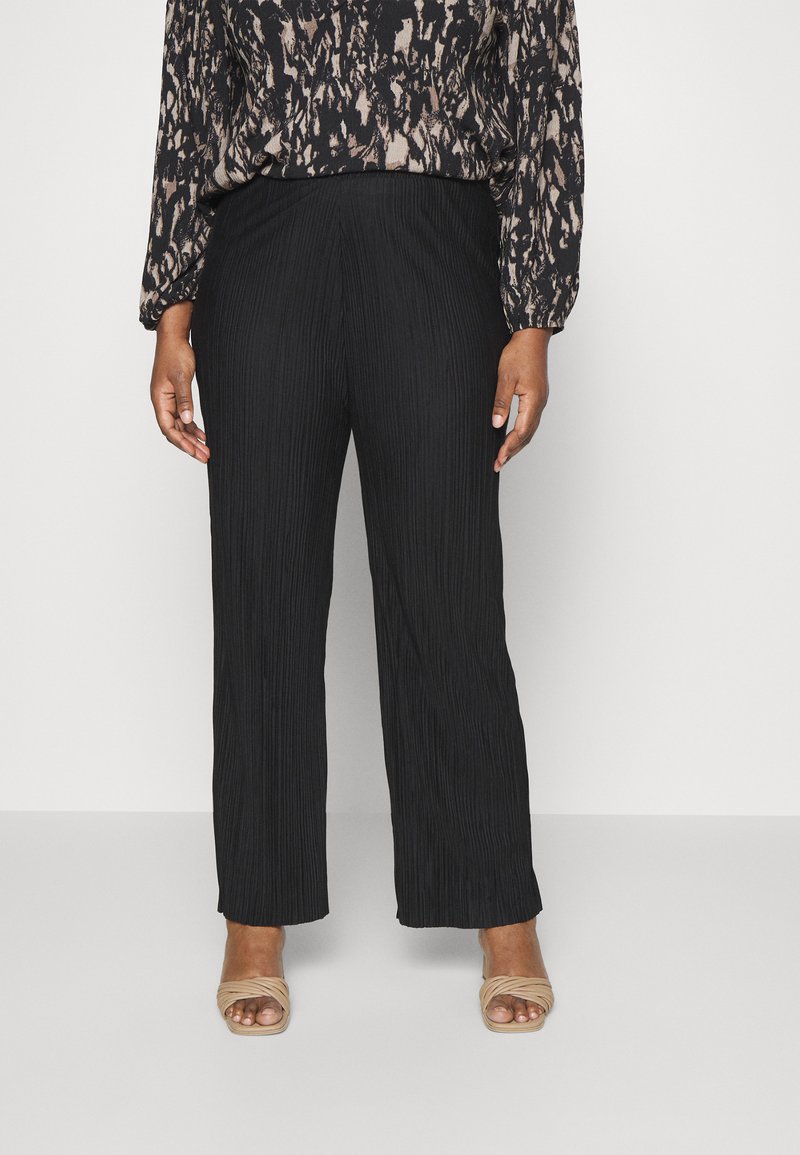 Pantalones plisados negros de cintura alta y corte holgado, combinados con una blusa estampada. La modelo lleva zapatos de tacón abiertos color beige.