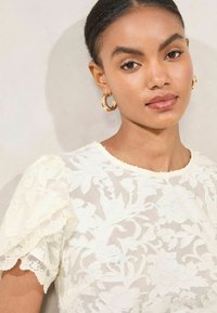 Floral-patterned, ivory blouse with sheer fabric, lace trim on short sleeves, crew neckline, and delicate texture. Gold hoop earrings.