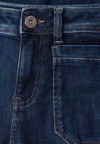 Tmavě modré džíny z denimu mají přední kapsu, kovový knoflík a kontrastní žluté švy. Textura vypadá hladce s mírným leskem.