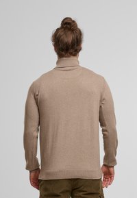 Mann mit braunen Haaren, die zu einem Dutt gebunden sind, trägt einen beigen Rollkragenpullover und olivgrüne Hose und steht vor einem schlichten hellgrauen Hintergrund.