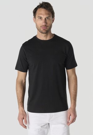 Hombre con el pelo corto y barba clara que lleva una camiseta negra lisa de manga corta y pantalones blancos, de pie frente a un fondo gris.