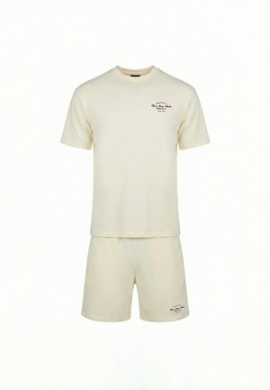 Mężczyzna noszący kremowy, oversize T-shirt z różowym napisem i pasujące krótkie spodenki, stoi tyłem na białym tle.