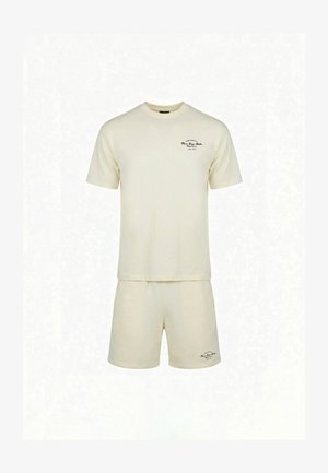 Muž nosiaci krémové oversized tričko s ružovým textom a zodpovedajúce šortky, stojaci otočený chrbtom na bielom pozadí.