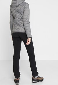 Veste isolante grise avec capuche, présentant un design matelassé et des coutures diagonales. Un pantalon noir et des bottes de randonnée marron complètent la tenue.