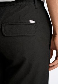 Zwarte broek met een achterzak, stofstructuur zichtbaar, met een klein logo-label. De tailleband lijkt elastisch te zijn.